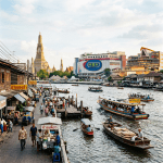 River in Bangkok with boats, people walking by market stalls, and Wat Arun temple